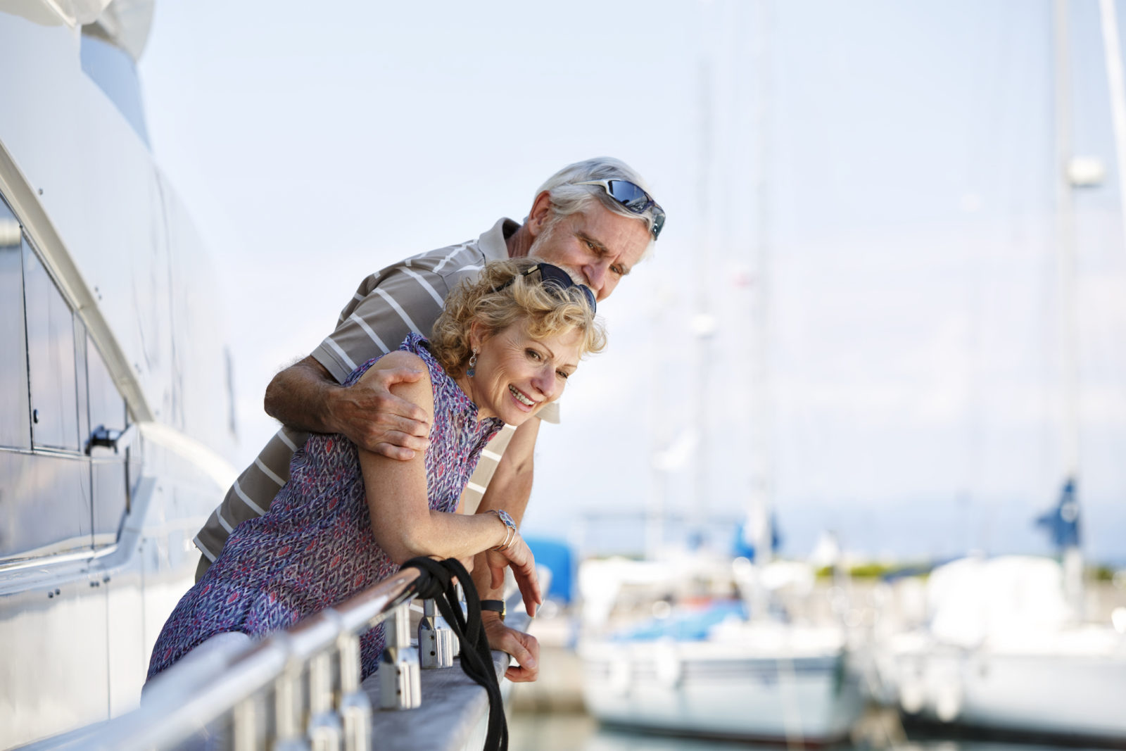 senior couple relaxing and sailing on their boat. The man is hugging his wife and they are smiling. In the background we can see the sea and some boats and yachts.