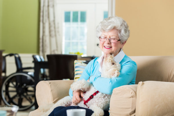 elderly woman in assisted living community in overstuffed chair with dog in her lap. Dog is giving the woman kisses and she is smiling