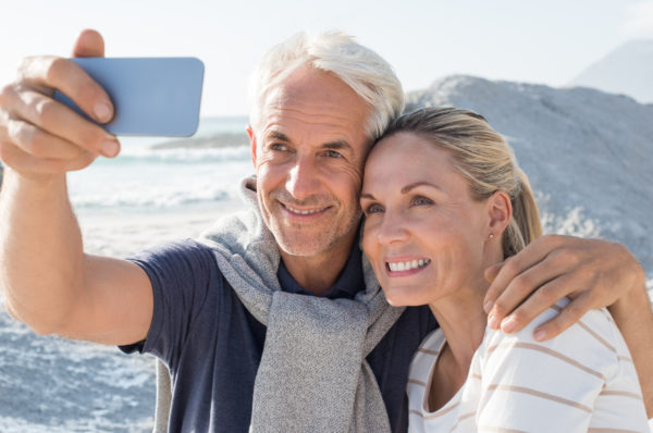 Happy romantic elder couple embracing on the beach and taking a photo with smartphone.