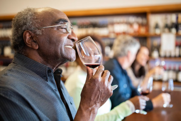 Close up of a senior Black man at wine tasting with people sitting at the back in tasting room. Man smelling wine with eyes closed.