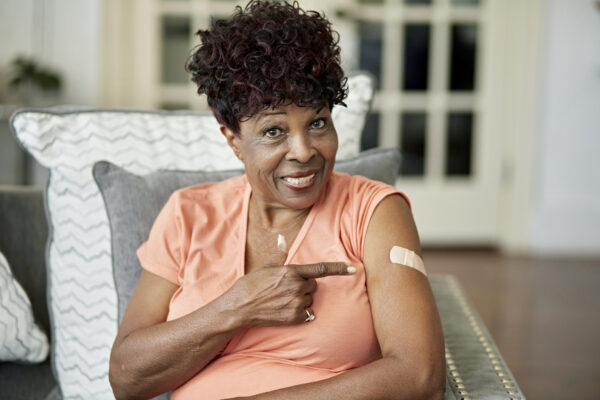 Waist-up view of casually dressed Black woman in late 70s sitting in family home and smiling at camera as she gestures with pride at adhesive bandage.