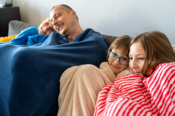 Father and daughters sitting on a sofa, cozying up under blankets.