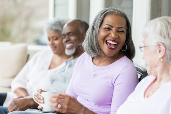A laughing African American senior woman sits on a front porch and drinks coffee with her friends.